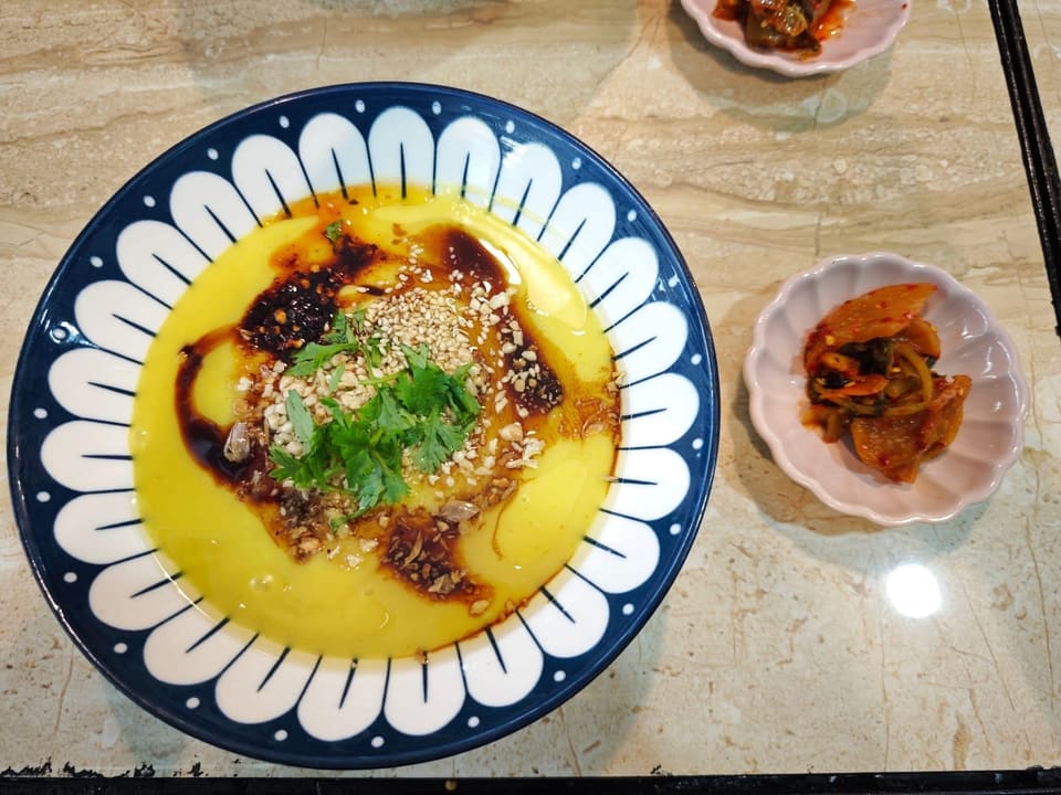 A bowl of warm yellow split-pea tofu, dressed with chilli oil and dark sauce, crushed peanuts, sesame seeds and fresh coriander, with a small dish of pickles beside it.