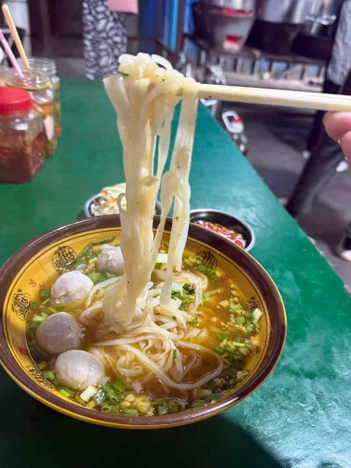 A bowl of noodle soup, the flat rice noodles lifted clear on chopsticks. Meatballs, chopped spring onion and coriander sit in a clear amber broth.