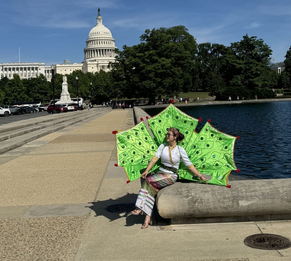 Ying Jein in Shan dance costume beside the reflecting pool in Washington DC, the Capitol dome behind her, wearing a pair of large green peacock-feather wings.