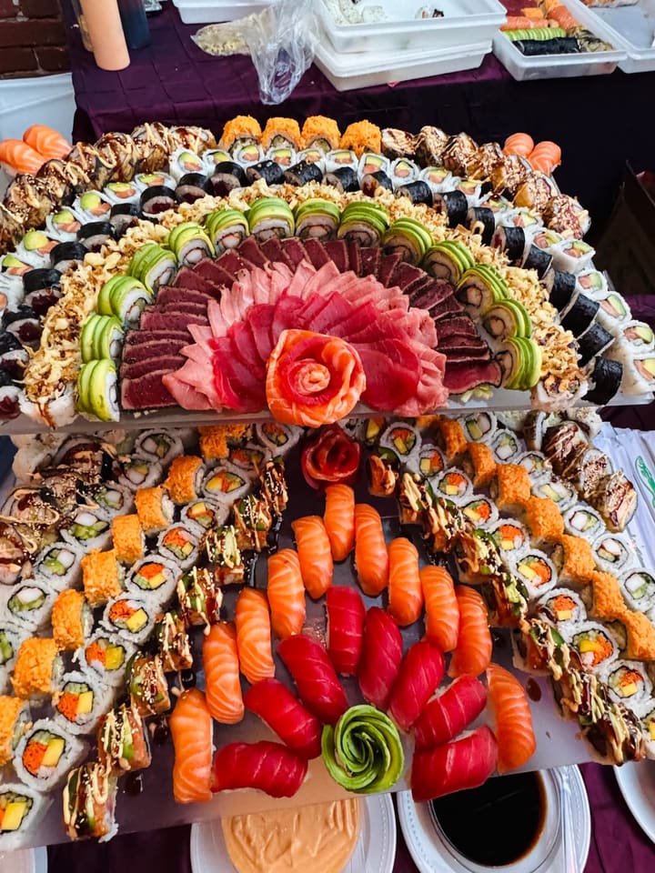 Two tiers seen from above: a fan of tuna and salmon sashimi around a salmon rose, framed by rows of avocado-draped and crisp-topped rolls.