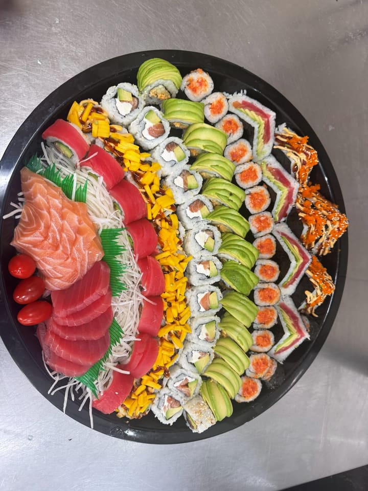 A round sushi tray seen from above: fanned salmon and tuna sashimi on shredded daikon, mango-topped and avocado-draped rolls, and spicy rolls finished with roe.